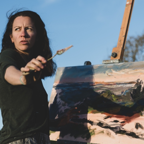 Women artist holding a paint brush stood in front of a picture on an easel