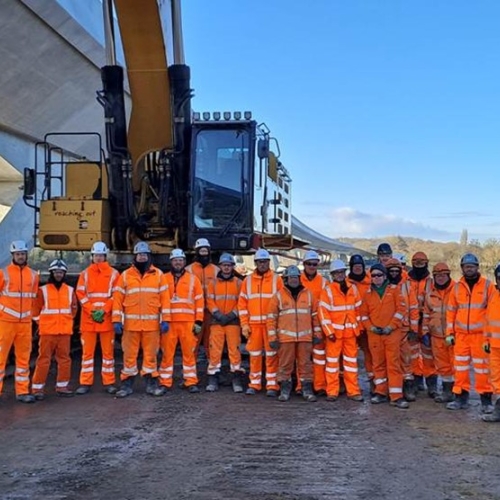 Group photo of men and women in orange hi vis workwear in front of a piece of construction equipment