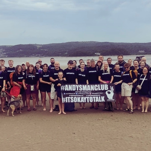 group of men and women stood together on a beach holding a black banner that says Andy's Man Club
