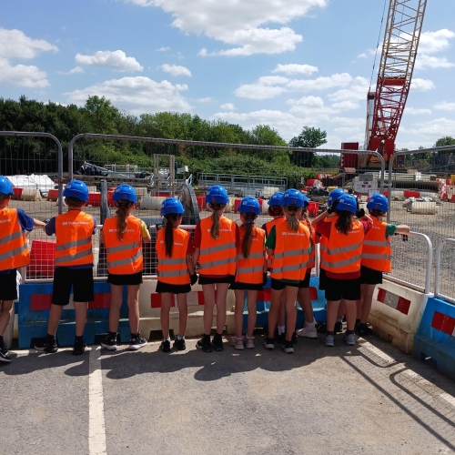 Group of school children in orange hi-vis wear and blue hard hats stood by fencing on a construction site