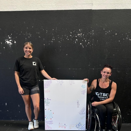 two women in gym wear holding a whiteboard, one is in a wheelchair