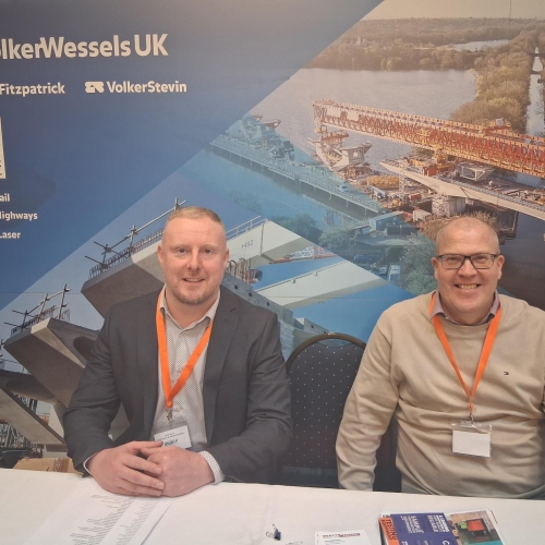 Two white men sat at a table in front of a blue banner with construction images on - VolkerStevin