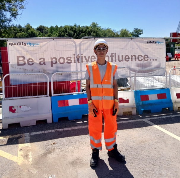 Young man wearing orange hi-vis workwear stood by fencing on a construction site