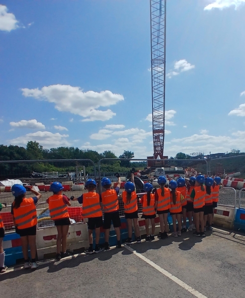 Group of school children in orange hi-vis wear and blue hard hats stood by fencing on a construction site