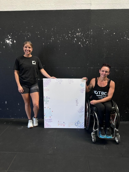 two women in gym wear holding a whiteboard, one is in a wheelchair