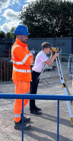 School boy stood looking through a surveying camera with a male in orange hi-vis construction wear and blue hard hat