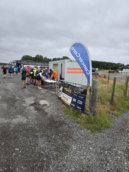 Cabin in a filed with Cancer Care logo on the side with a free standing banner. People stood outside the cabin registering at a table