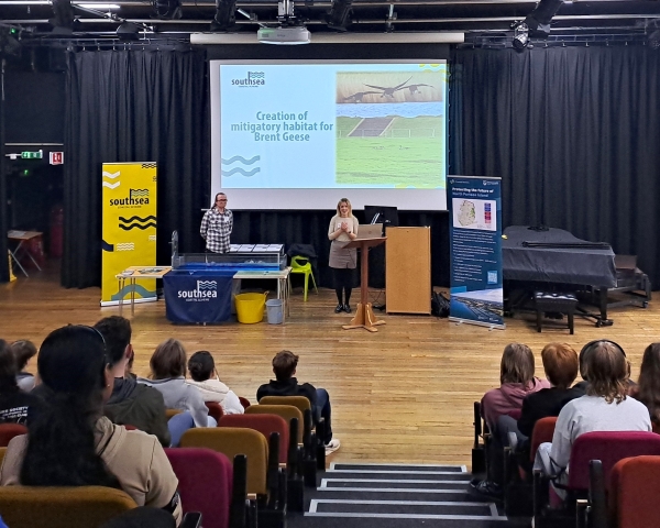 Women and man stood in an auditorium in front of a group of students and large screen with a presentation on it 