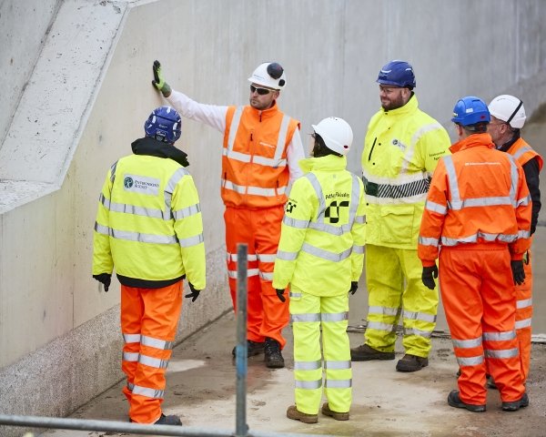 Men and woman in orange and yellow hi vis workwear stood in a circle in front of a concrete wall