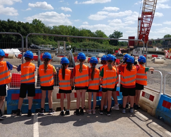 Group of school children in orange hi-vis wear and blue hard hats stood by fencing on a construction site