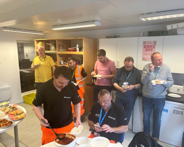 Group of men and women in a site cabin kitchen stood talking and eating buffet food