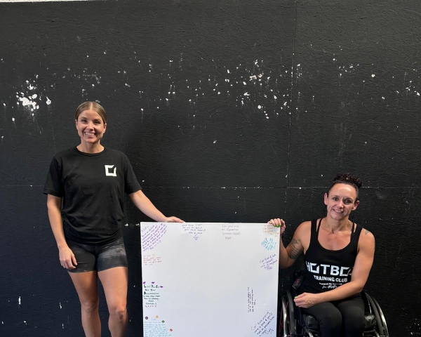 two women in gym wear holding a whiteboard, one is in a wheelchair