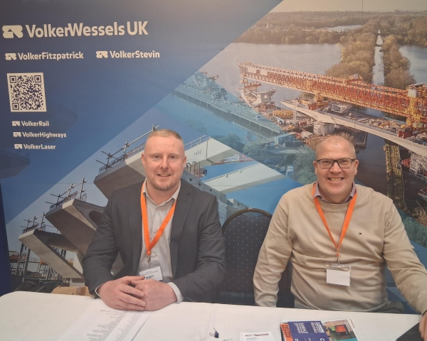 Two white men sat at a table in front of a blue banner with construction images on - VolkerStevin