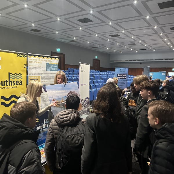 Two women stood behind a table with banners behind them, showing a group of children information on the Southsea flood scheme