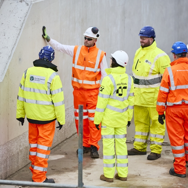 Men and woman in orange and yellow hi vis workwear stood in a circle in front of a concrete wall
