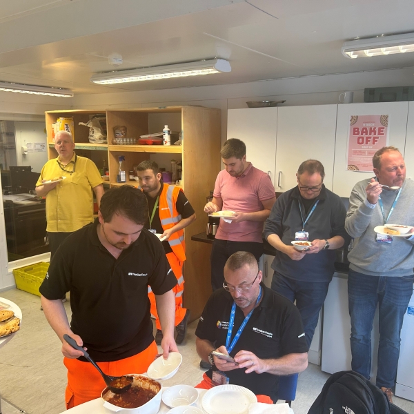 Group of men and women in a site cabin kitchen stood talking and eating buffet food