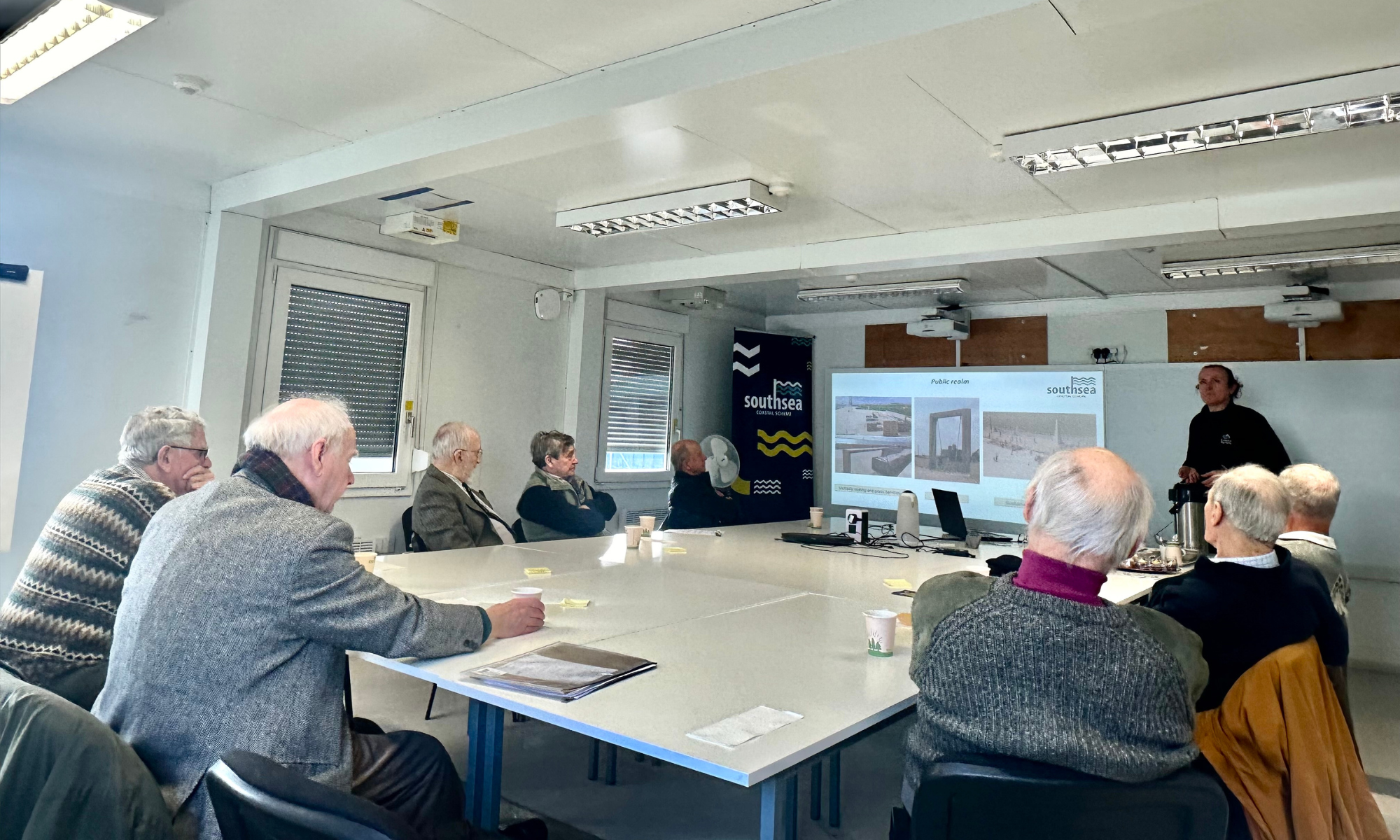 A group of male retired engineers sat round a table looking at a presentation on a big screen