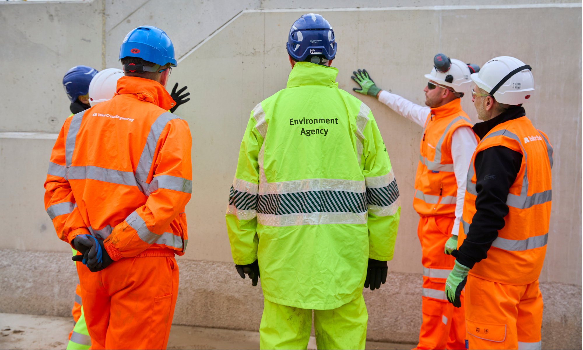 Men and woman in orange and yellow hi vis workwear stood in a circle in front of a concrete wall