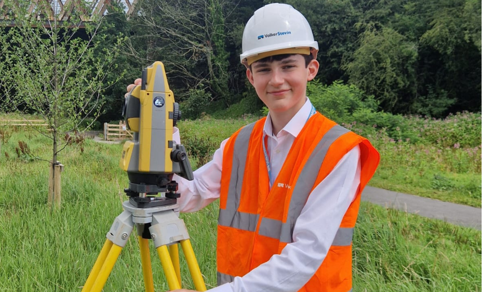 Young man with orange hi-vis vest and white hard hat stood by construction equipment