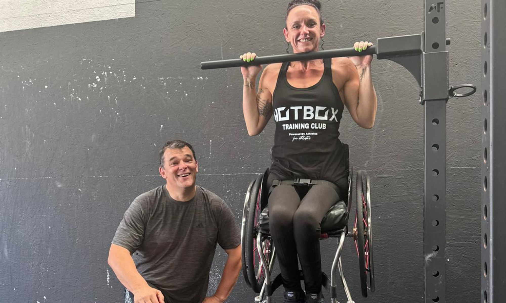 Women in a wheelchair lifting on a piece of gym equipment with a man in black gym wear stood next to her