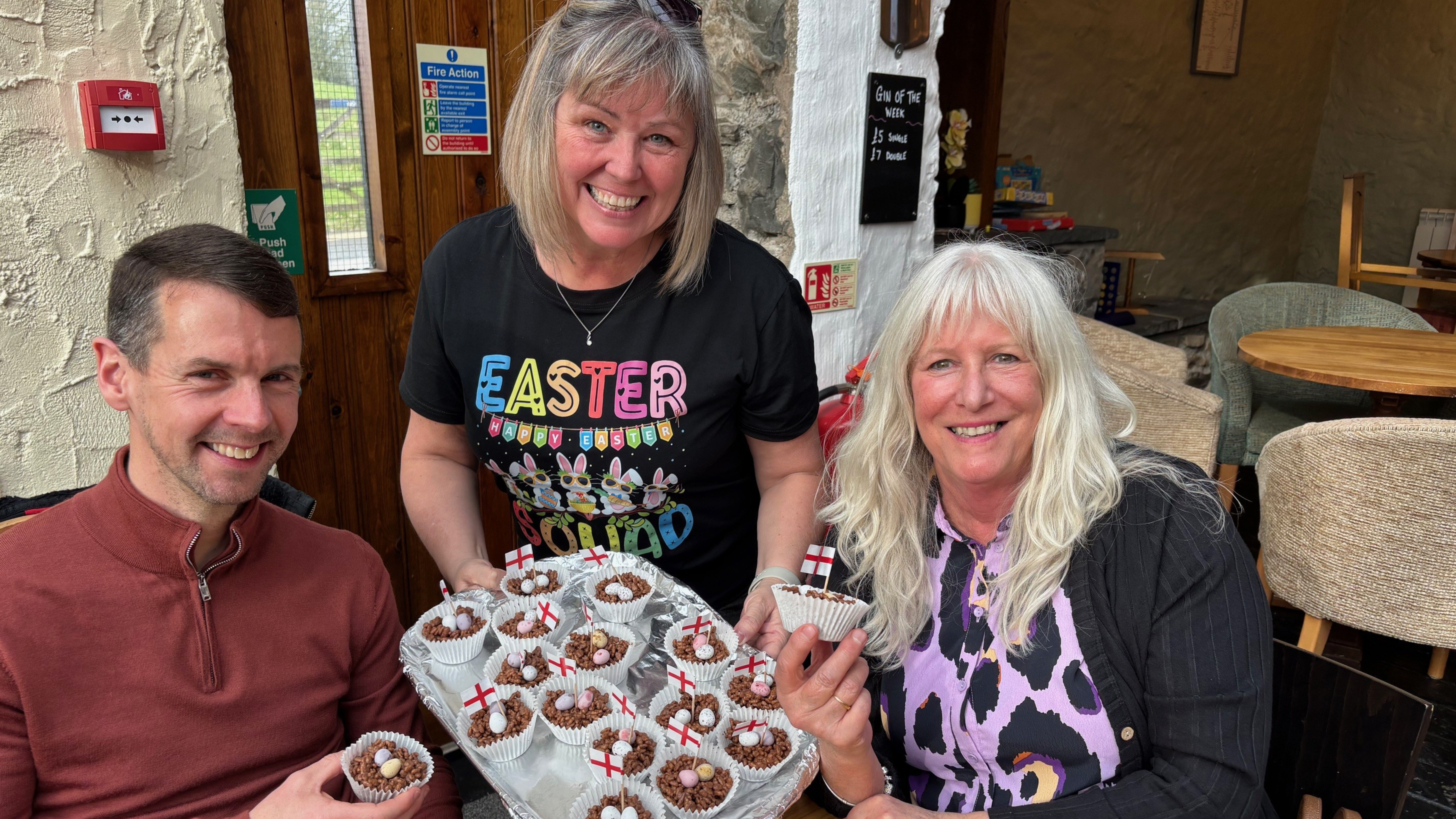 Two women and a man sat at a table holding cakes that have the St. George's flag on