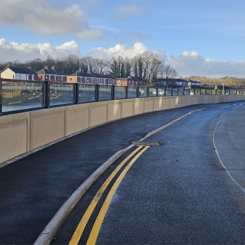 Landscape photo of main road and pavement alongside a concrete river flood wall with glass panels on top