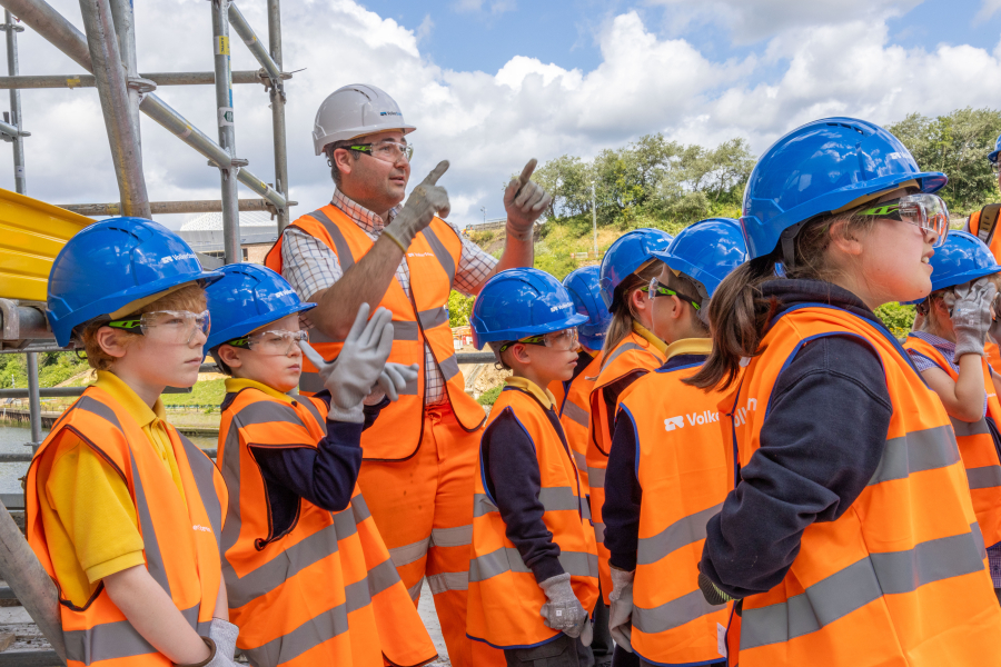 A man talking to a group of children on a construction stie all wearing orange hi-vis workwear and blue and white hard hats