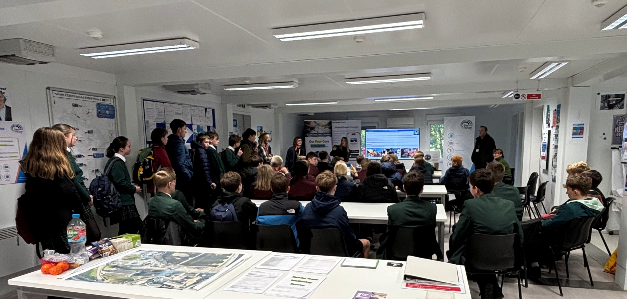 Group of school children and adults stood in a site cabin around a table and in front of a large screen