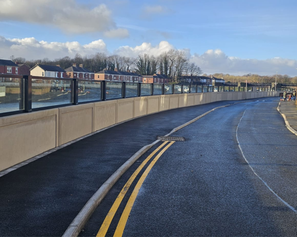 Landscape photo of main road and pavement alongside a concrete river flood wall with glass panels on top