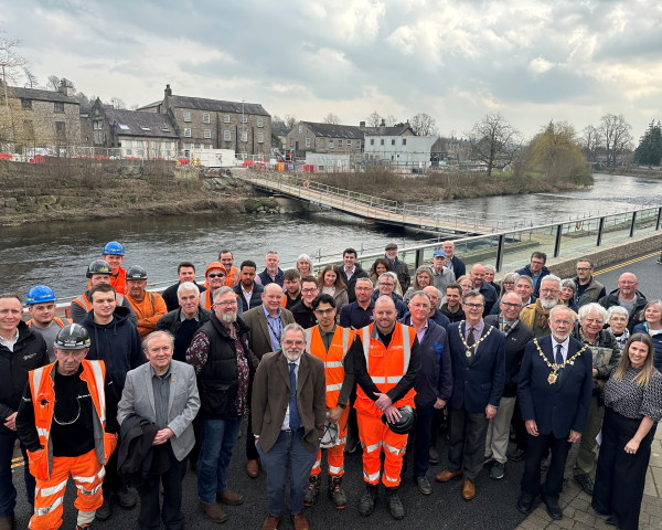 Group of men and women some in orange hi-vis workwear stood on a bridge with a river behind